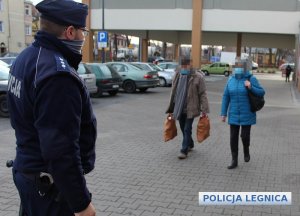 umundurowany policjant stoi na parkingu sklepu a w jego kierunku idzie kobieta i mężczyzna niosący torby z zakupami w tle po lewej stronie zaparkowane pojazdy