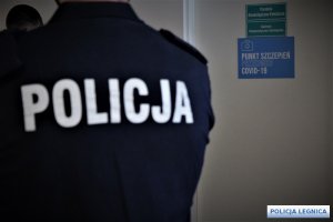 napis policja na mundurze policjanta stojącego przy drzwiach na którym jest kartka szczepienia przeciw covid-19