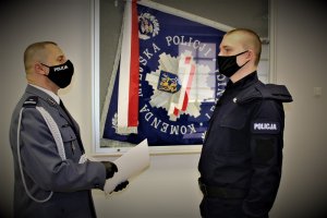 Komendant Miejski Policji w Legnicy wręcza policjantowi akt ślubowania. Funkcjonariusze stoją ubrani w policyjny mundur przy gablocie w której jest sztandar Komendy Miejskiej Policji w Legnicy