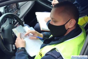 policjant siedzi w radiowozie na twarzy ma założoną maseczkę a w ręce trzyma notatnik i dowód rejestracyjny