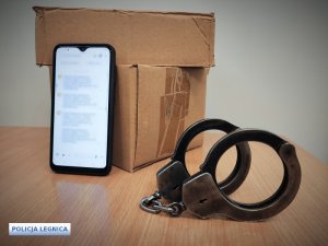 Dwa pudełka kartonowe ustawione jeden na drugim, a o nie oparty telefon komórkowy i kajdanki policyjne.