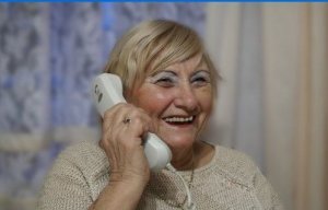 Seniorka trzyma słuchawkę telefonu przy uchu i się uśmiecha.