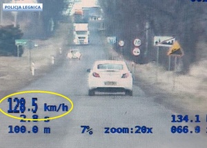 Widok kamery wideorejestratora przedstawiający drogę, na której poruszają się pojazdy. W lewym dolnym rogu zaznaczona na żółto prędkość poruszającego się samochodu koloru białego z wynikiem 128,5 km/h