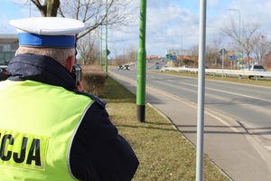 Policjant ruchu drogowego stoi przy drodze i trzyma ręczny miernik prędkości, a po drodze poruszają się pojazdy.