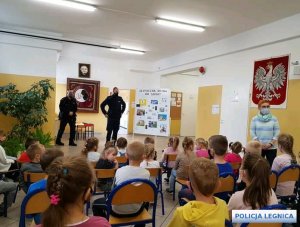 dwóch policjantów  stoi przodem do uczniów siedzących na krzesełkach i rozmawia z nimi o ich bezpieczeństwie