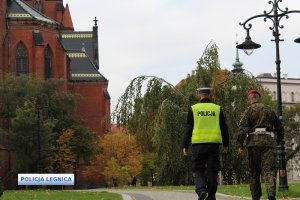 Policjant wspólnie z funkcjonariuszem Żandarmerii Wojskowej idą skwerem, po prawej stronie latarnia a po lewej katedra, po obu stronach rosnące drzewa