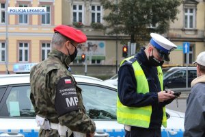Policjant legitymuje obywatela, a lewej strony stoi funkcjonariusz Żandarmerii Wojskowej za nimi radiowóz a w tle kamienica i rosnące drzewo