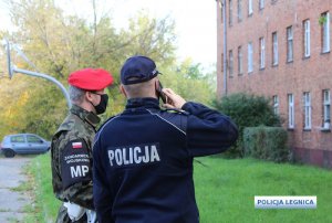 policjant trzyma telefon przy uchu obok stoi funkcjonariusz Żandarmerii Wojskowej a po prawej stronie budynek mieszkalny wielorodzinny. Między mundurowymi a budynkiem pas zieleni.