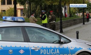 na pierwszym planie bok radiowozu a w tle policjant z funkcjonariuszem Żandarmerii Wojskowej w maseczkach stoją przy skwerze i legitymują mężczyznę w oddali po prawej stronie idzie kobieta