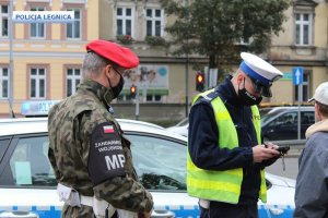 Policjant ruchu drogowego legitymuje mężczyznę na ulicy przy radiowozie, obok stoi funkcjonariusz Żandarmerii Wojskowej w tle kamienica