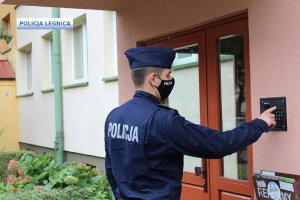 policjant stoi przed wejściem do klatki schodowej i trzyma palec na nr 1 dzwoniąc domofonem