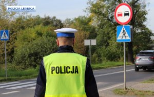 policjant ruchu drogowego stoi przed oznakowanym przejściem dla pieszych, po prawej stronie znak zakazu wyprzedzania a w oddali samochód