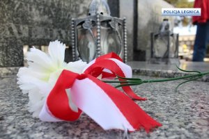 na pomniku leży biały goździk z zawiązaną wstążką w kolorze biało-czerwonym, za nim stoi znicz a w oddali po prawej stronie idzie mężczyzna