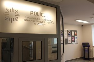 Poczekalnia z przeszklonym okienkiem recepcyjnym, na którym widnieje napis  stanowisko kierowania komendy miejskiej policji w Legnicy na ścianie tablica z zmieszczonymi ogłoszeniami i plakatami oraz infomat