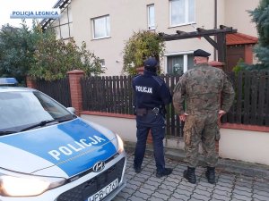 Policjant z żołnierzem stoją na chodniku przed ogrodzeniem przy wejściu na posesję przed domem, z lewej strony zaparkowany radiowóz