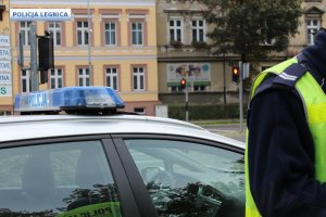 policjant ruchu drogowego stoi przy radiowozie a w tle kamienica i sygnalizator świetlny nadający sygnał czerwony dla pieszych