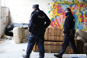 dwóch policjantów stoi w pomieszczeniu w którym są tapczany oraz fotel a na ścianie są grafiti